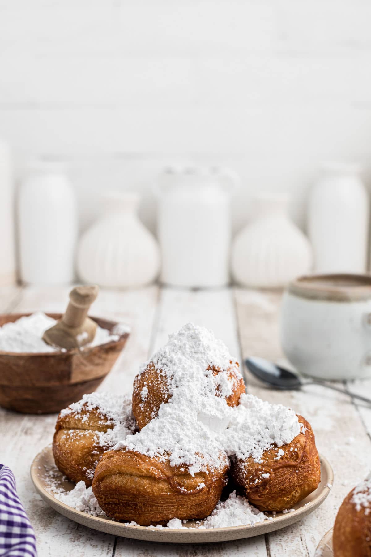 a plate of biscuit beignets with heaps of powdered sugar on top, with a coffee in the background