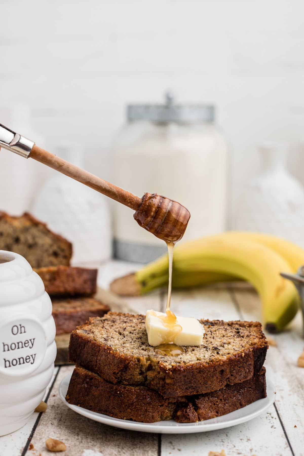 2 slices of banana bread with a butter slice on top with honey being drizzled on top - bananas in the background