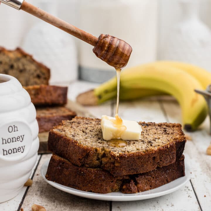 2 slices of banana bread with a butter slice on top with honey being drizzled on top - bananas in the background
