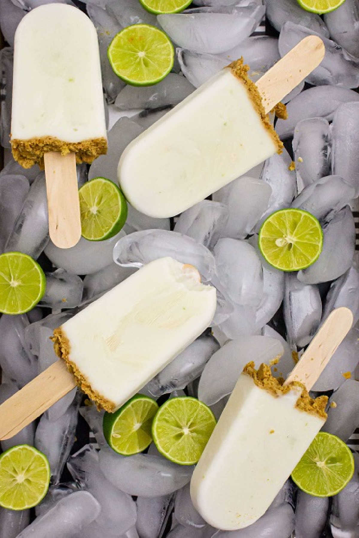 key lime pie popsicles laying on top of ice with slices of limes scattered around and one with a bite taken out of it