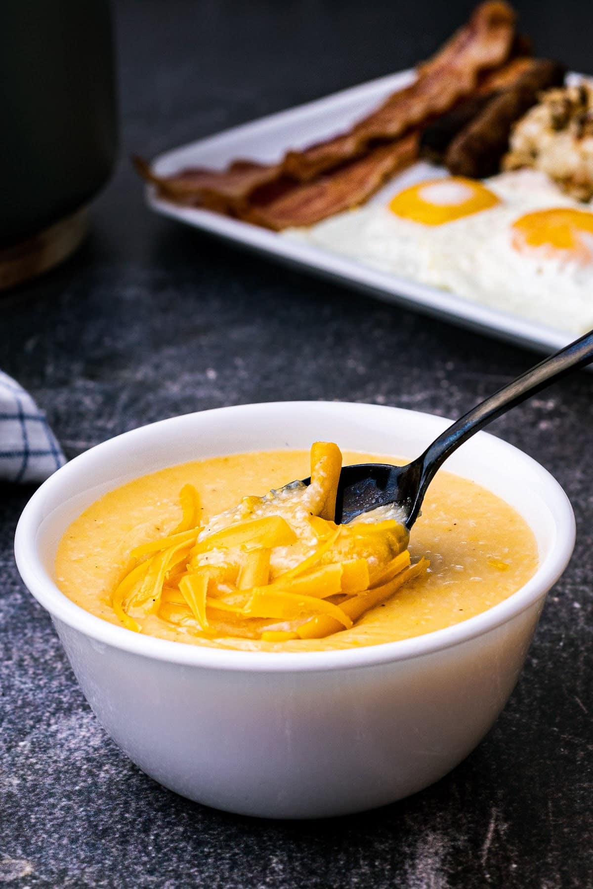 a bowl of cheese grits with a spoon taking some with some bacon eggs in the background