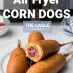 a plate with corn dogs piled, the top one having a bite taken out with an air fryer in the back ground