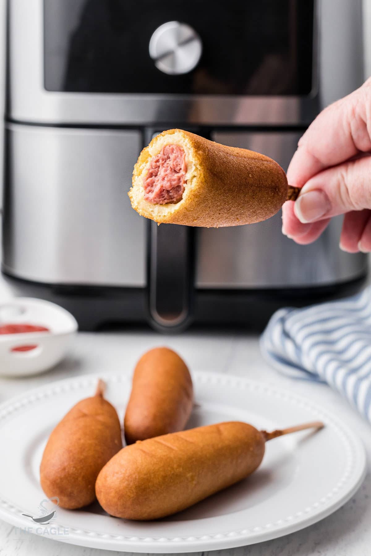 a hand holding a corn dog that has a bite taken out of it in front of an air fryer