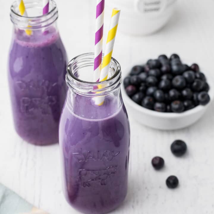 a purple looking drink inside two milk bottles, this is blueberry milk. With a bowl of blueberries in the background