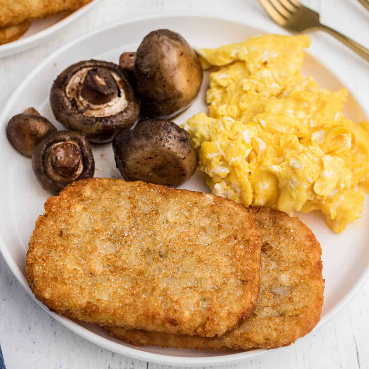 plate with 2 hash brown patties on it, with scrambled eggs and mushrooms in the back