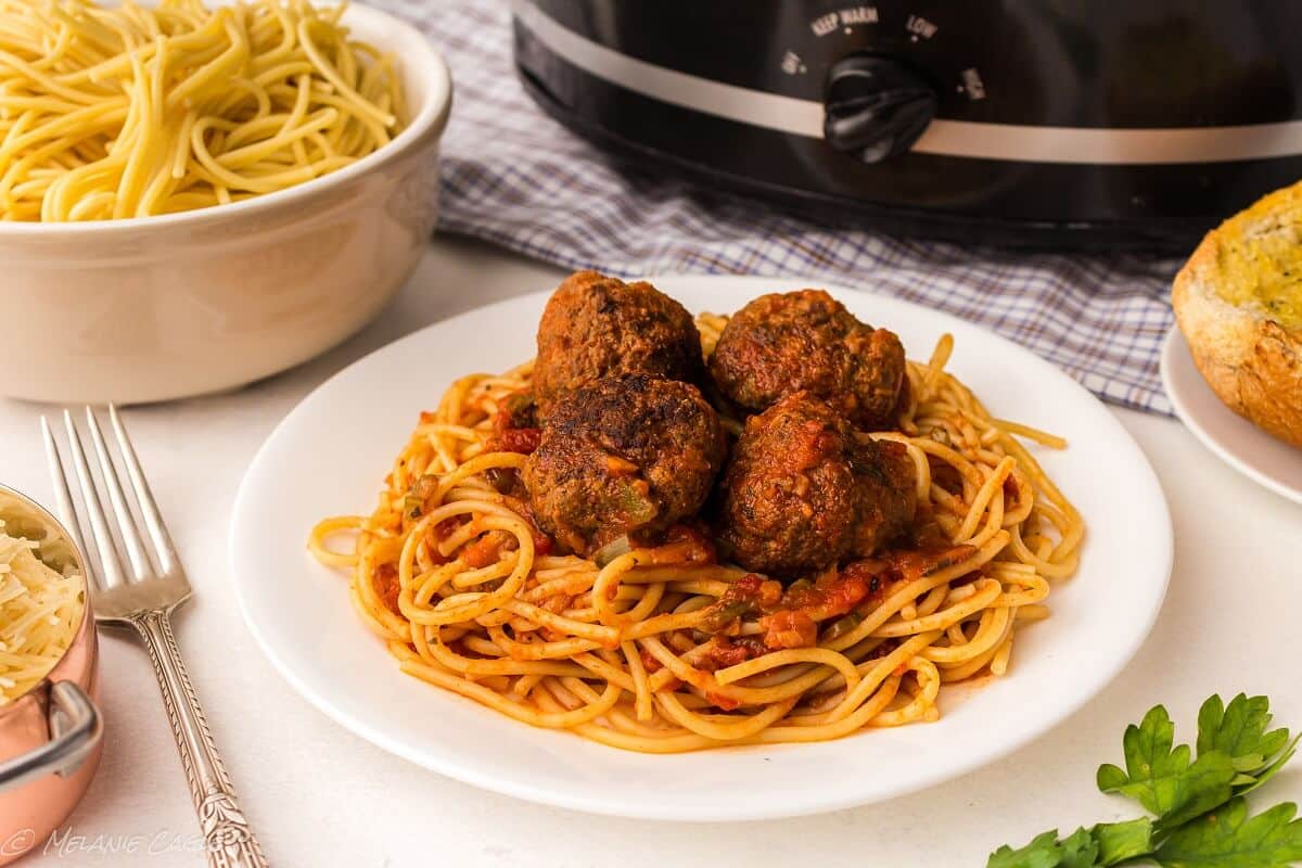 a plate of spaghetti with four meatballs on top - a crock pot in the back