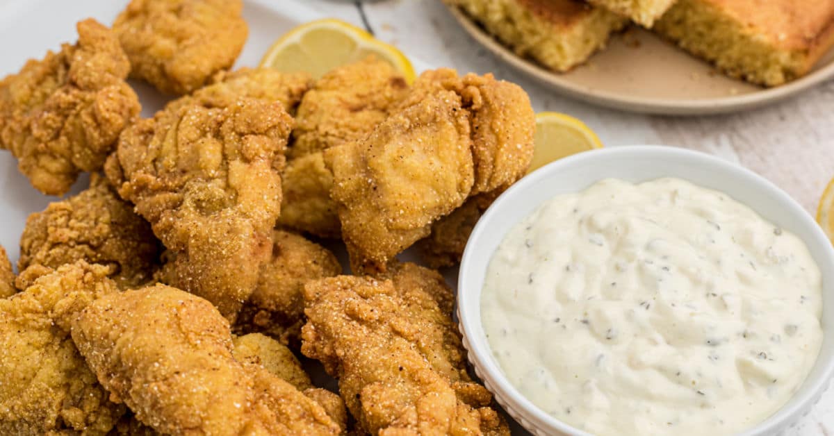 close up of some fried catfish nuggets with some tartar sauce and cornbread in the background