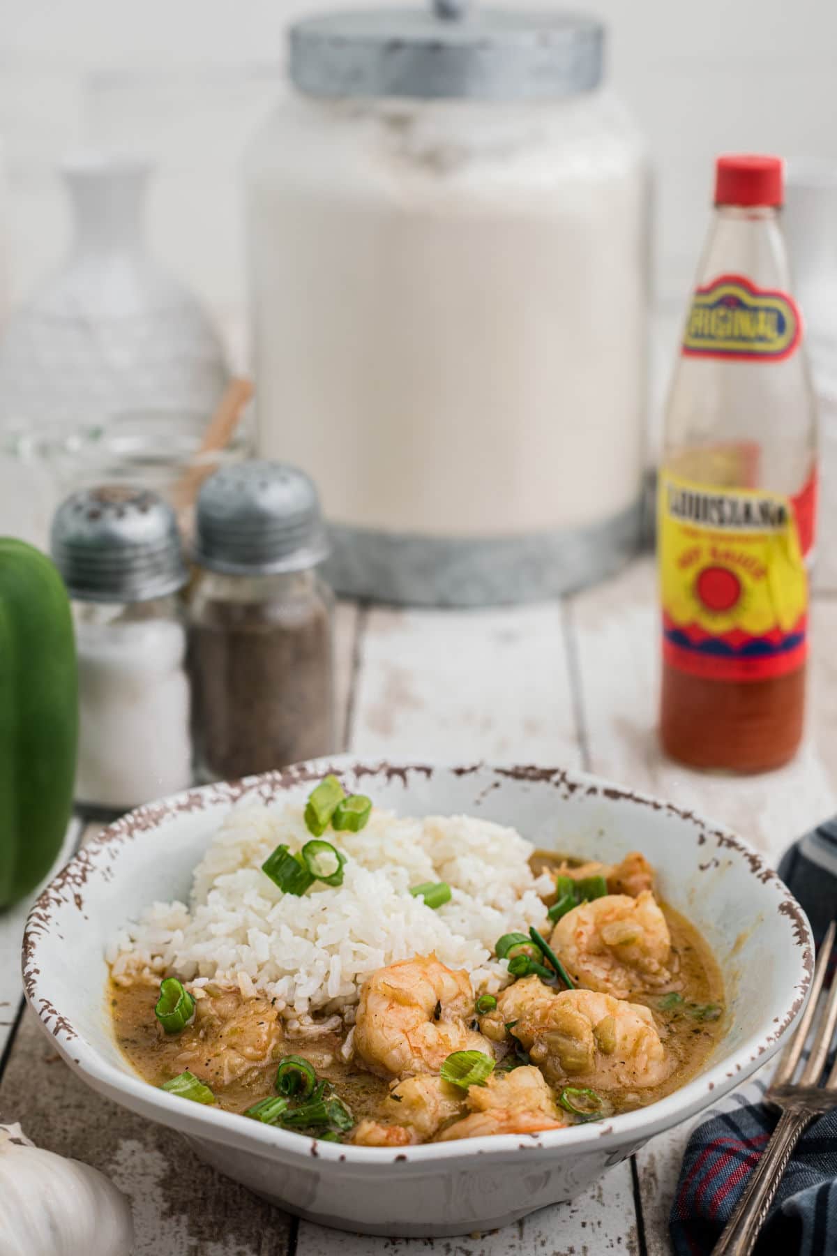a bowl full of shrimp etouffee with a pile of rice and garnished with chopped green onions with a bottle of hot sauce in the background