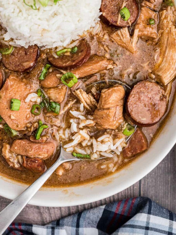 a close up of a bowl of chicken and sausage gumbo with a spoon sitting in it, looks like it's about to lift up, with some rice too