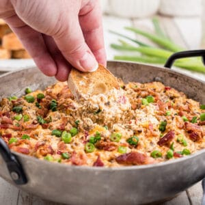 a skillet with some boudin dip cooked and some green onions sprinkled on top with a hand dipping into the dip