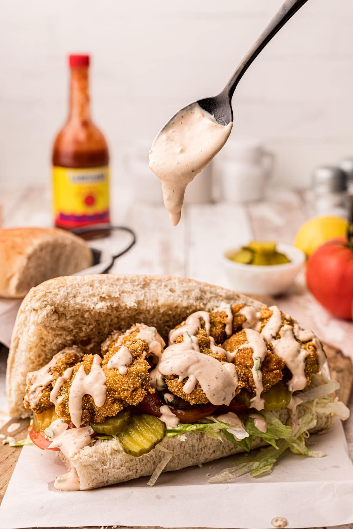 a fried oyster po'boy with remoulade sauce being drizzled onto it from above with some hot sauce in the background