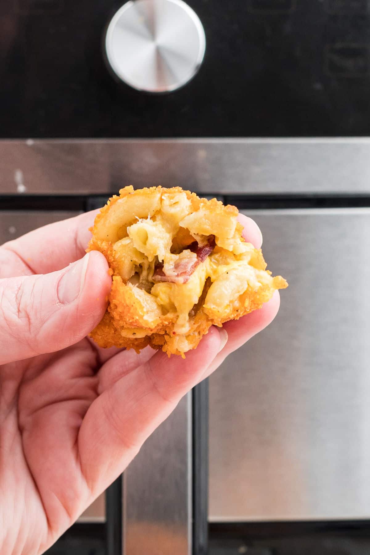 A hand holding a mac and cheese bite in front of an air fryer.