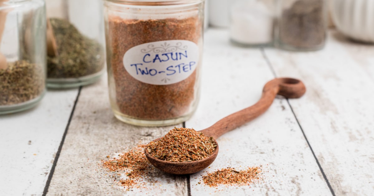 A jar of Cajun two step seasoning behind a wooden spoon piled high with the seasoning.