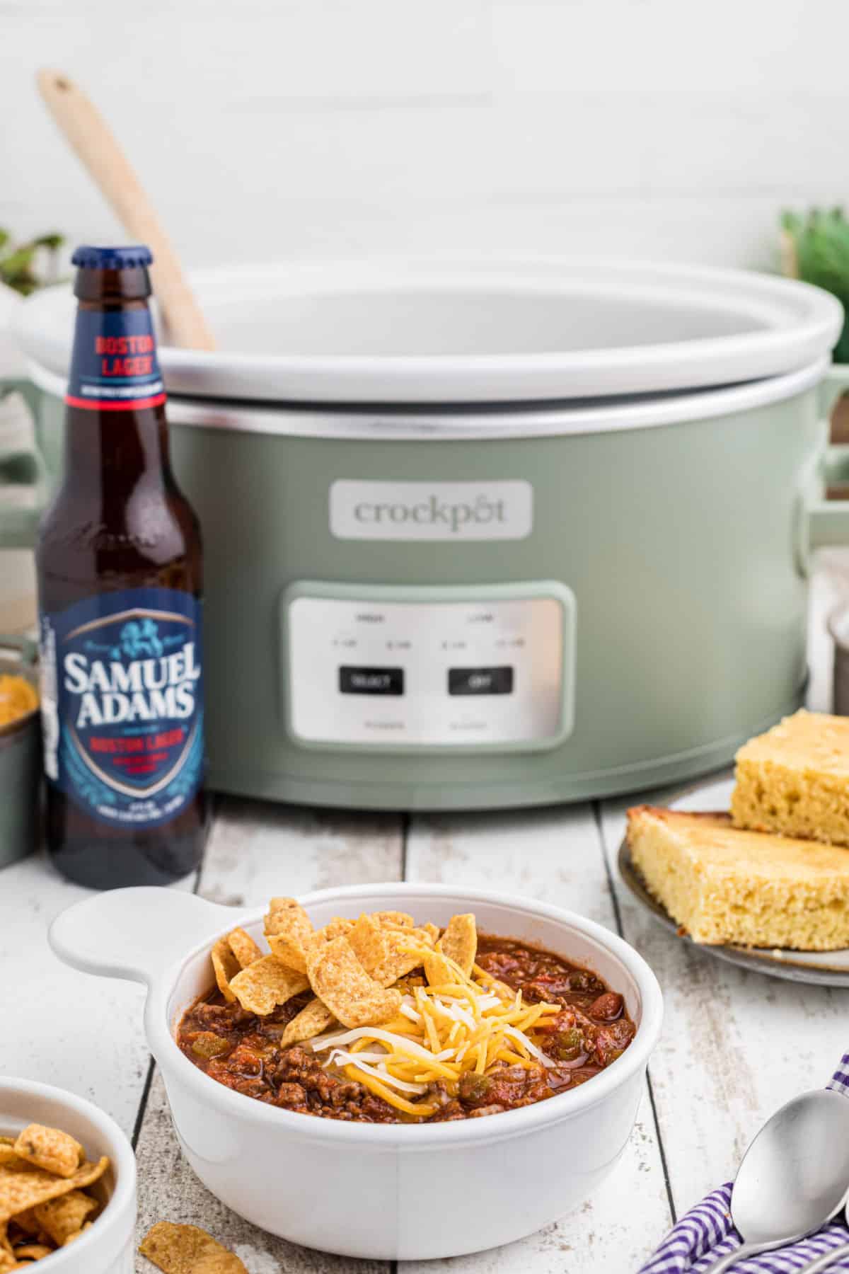 A dished out bowl of chili in front of a crock pot with a bottle of beer stood next to it.