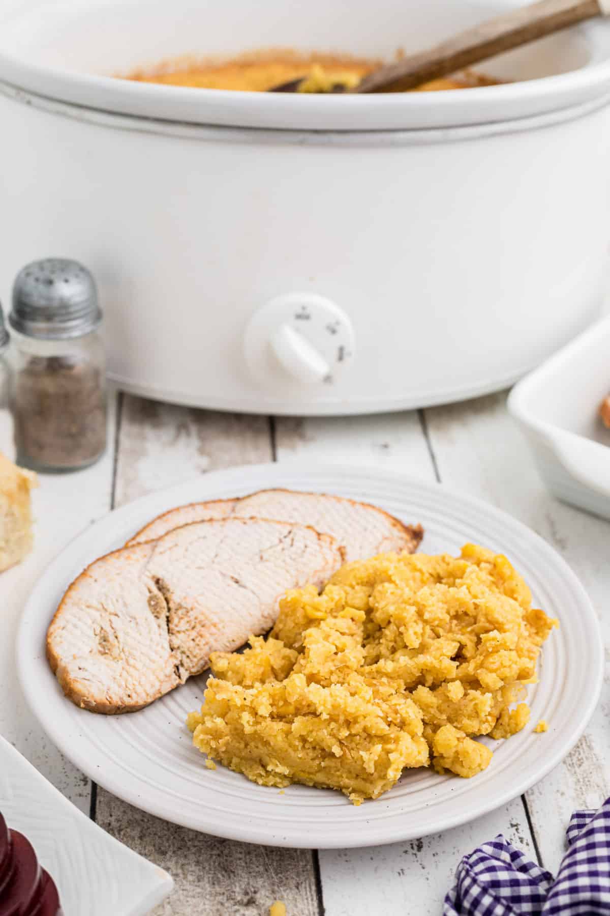 A plate of corn casserole with some slices of turkey behind with a crock pot.