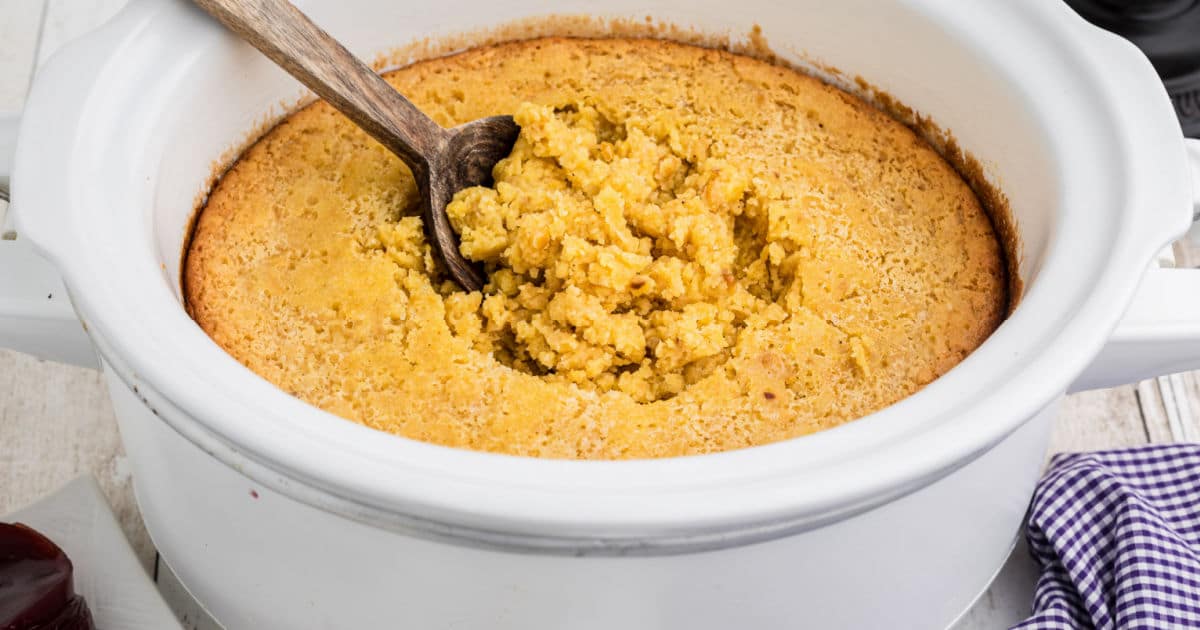 a corn casserole in a slow cooker with a wooden spoon digging into it.