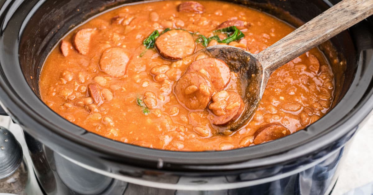 a pot full of beans with sausage slices and a spoon digging in.