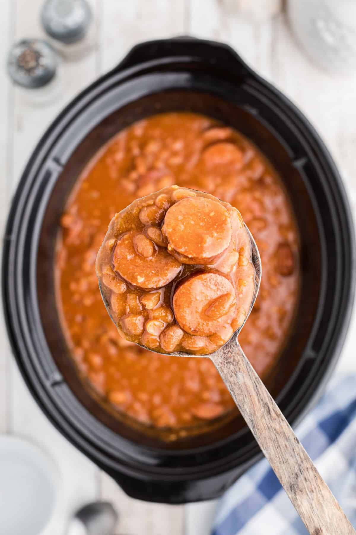 Overhead shot of a spoon holding some kielbasa and baked beans over a slow cooker in the background.