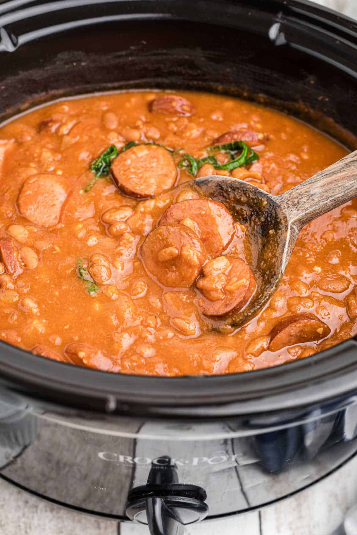 Close up side view of a crock pot with kielbasa and baked beans with a spoon digging in.