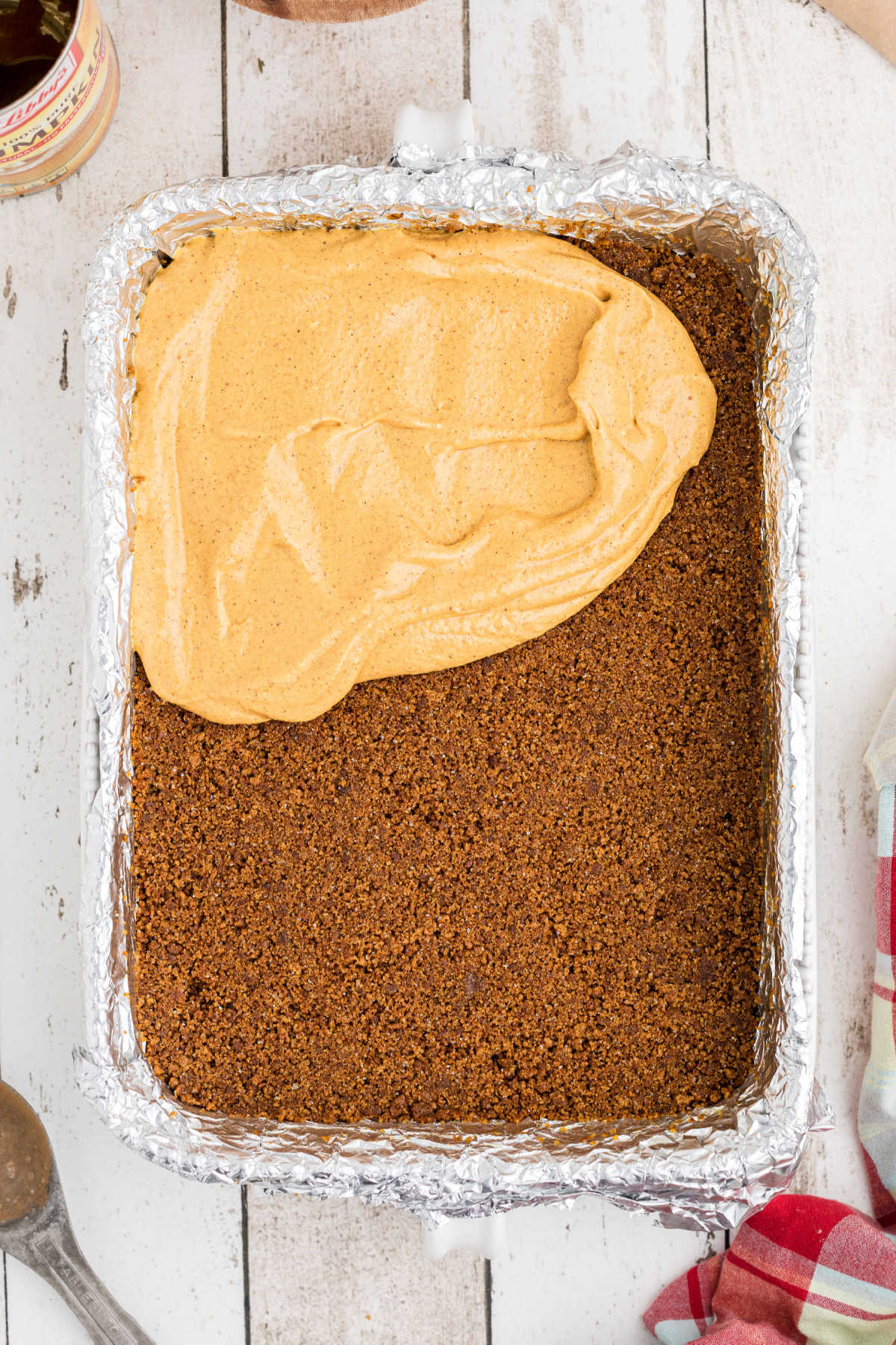 A large baking dish with a cookie crumb base, with a pumpkin cheesecake filling being spread on top.