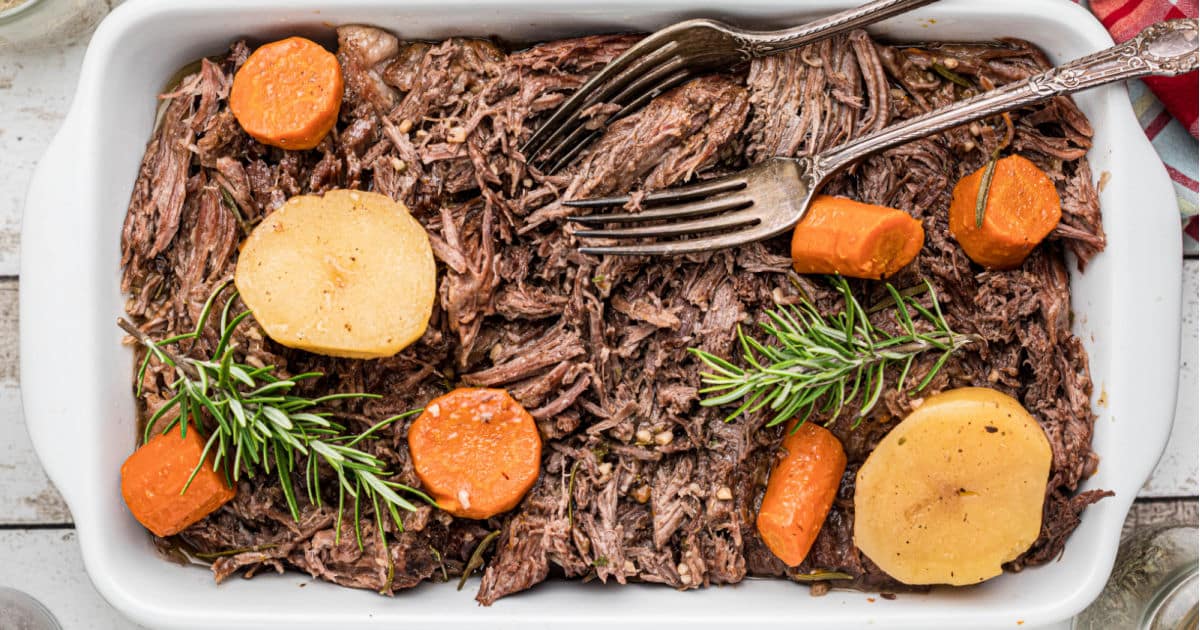 a dish full of tri tip roast that has been shredded with forks with potatoes and carrots.
