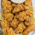A large platter of fried catfish nuggets with some lemon wedges and a tartar sauce.
