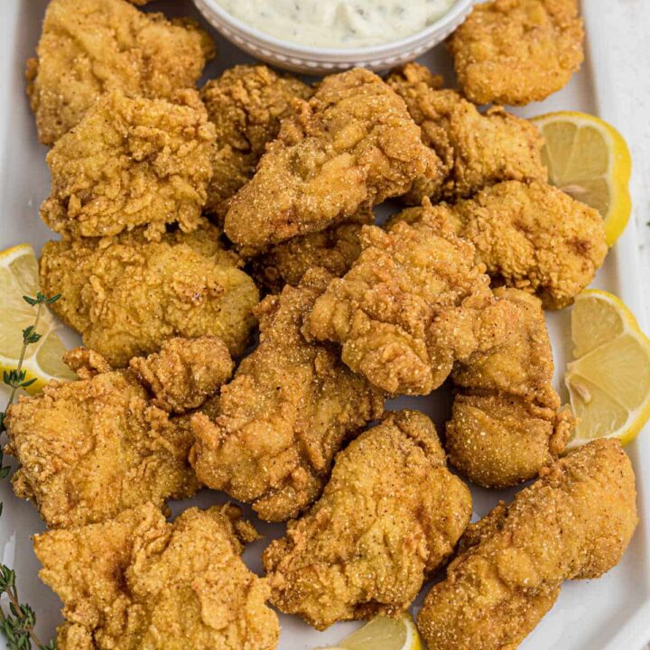 A large platter of fried catfish nuggets with some lemon wedges and a tartar sauce.