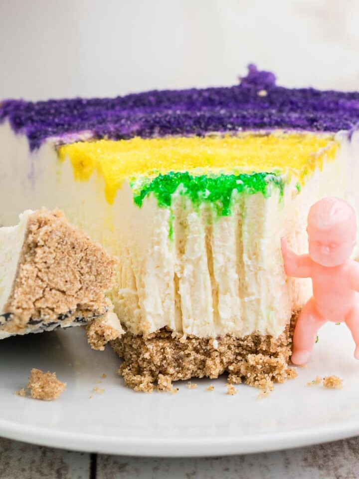 Close up of a slice of Mardi Gras cheesecake with a king cake baby on the side.
