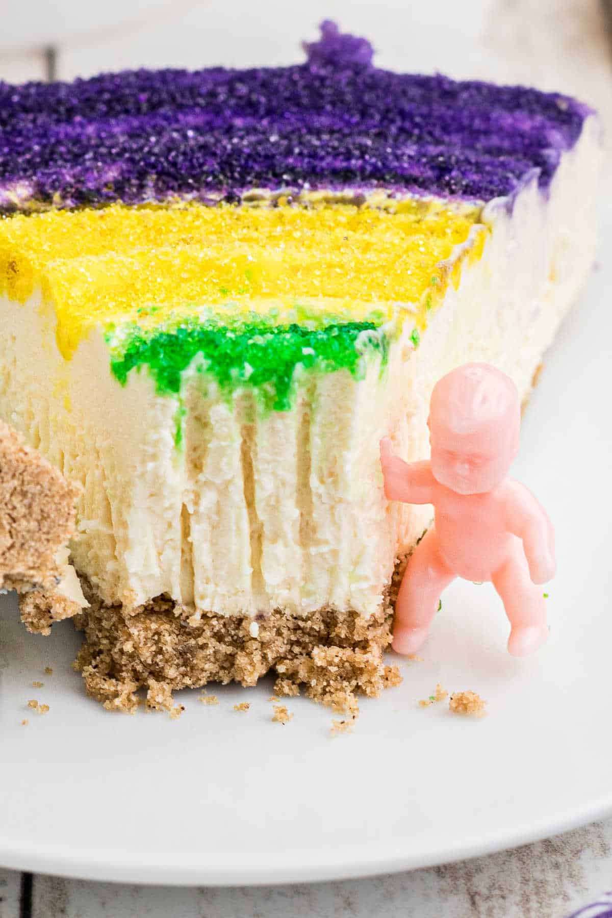 Close up of a slice of mardi gras cheesecake with a king cake baby on the side.