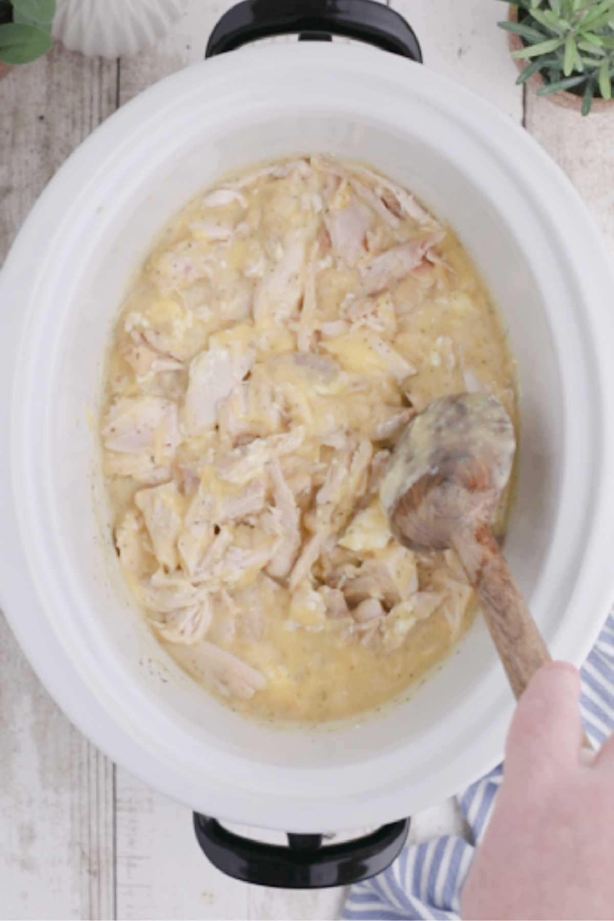 Shredded chicken added to a crock pot with other ingredients and being stirred together.