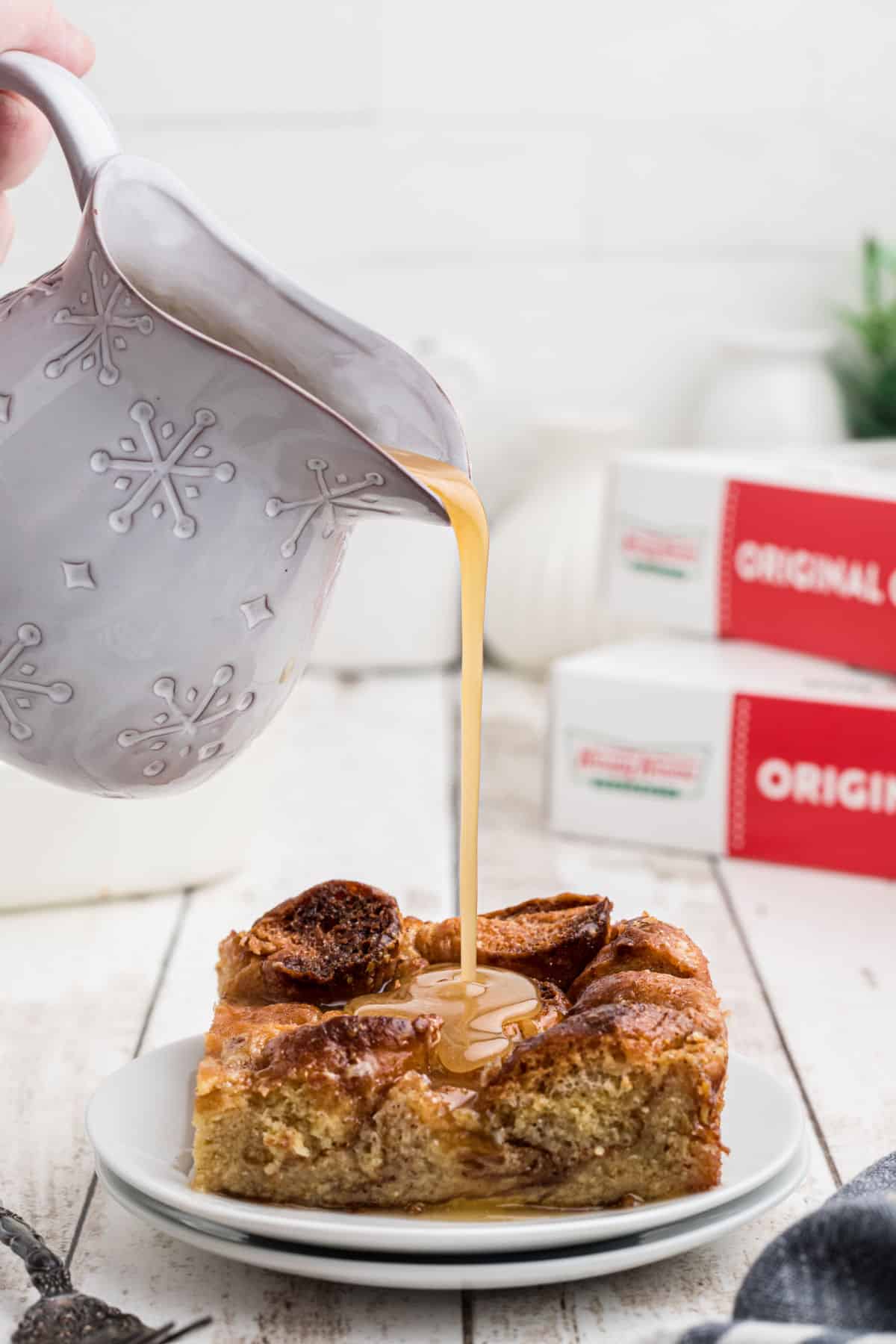 A slice of krispy kreme bread pudding on a plate with some vanilla sauce being poured over the top.