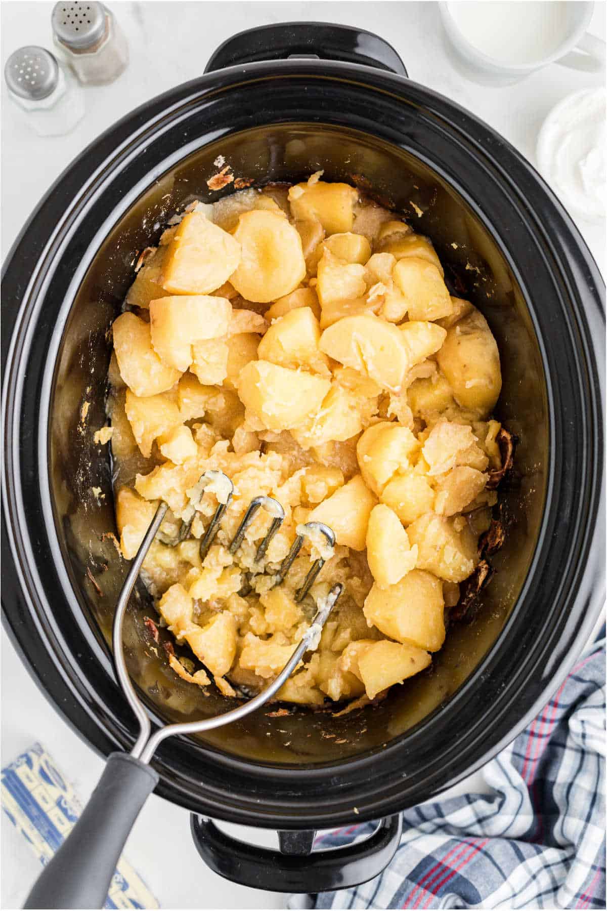 Potatoes being mashed with a potato masher in a slow cooker.