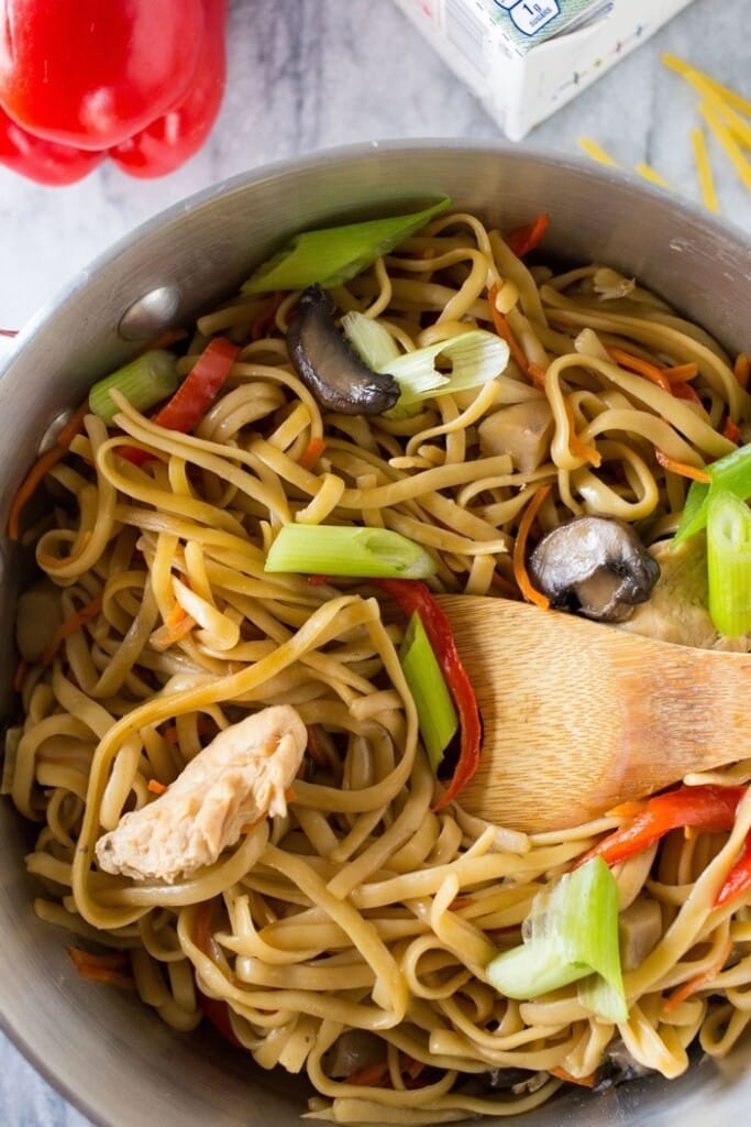 A pan with Chinese Lo Mein noodles and vegetables, with a wooden spoon stirring it.