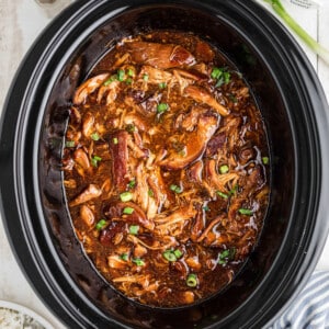 Overhead shot of a crock pot with bourbon chicken inside sprinkled with green onions.
