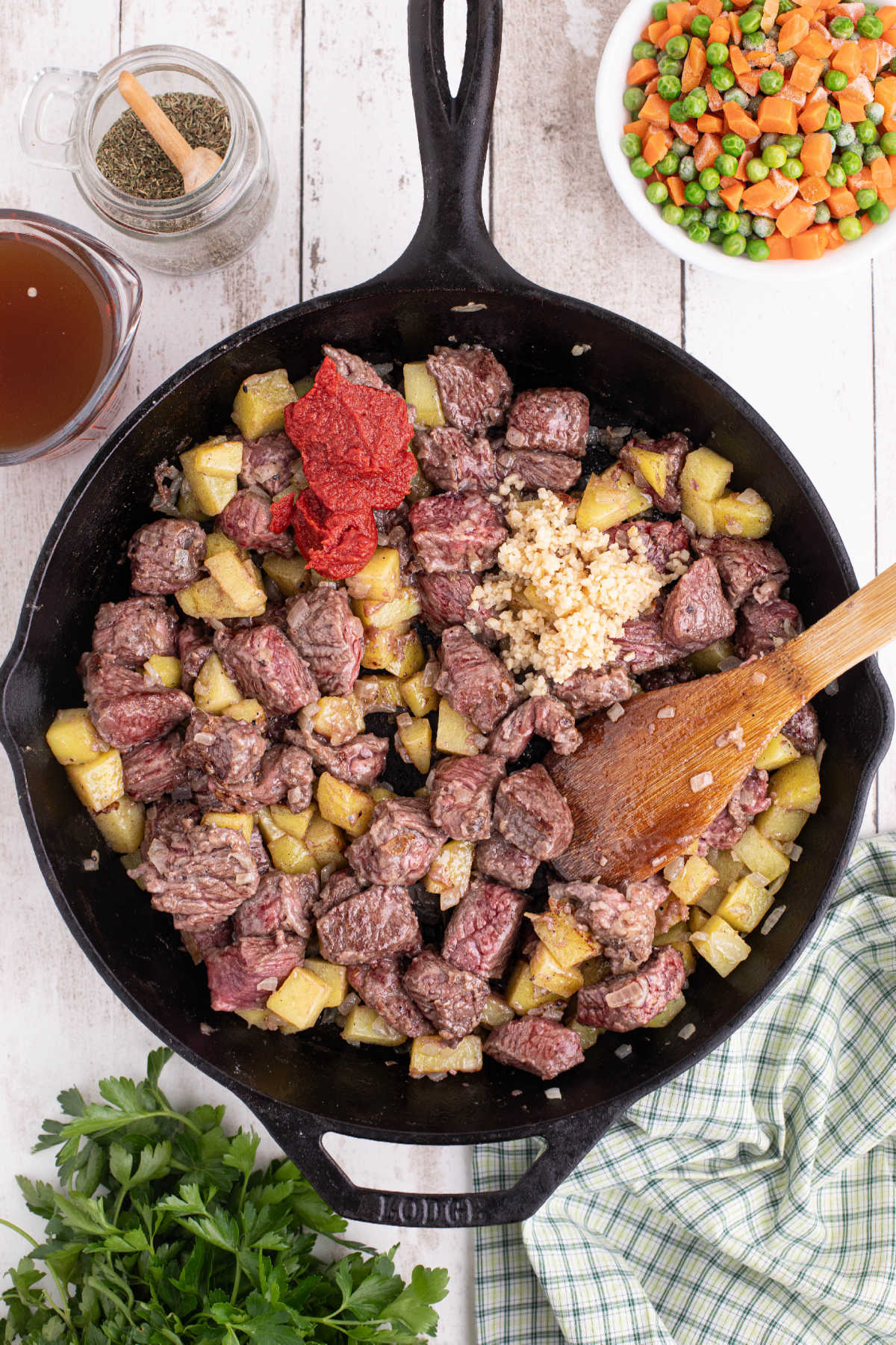 A large cast iron skillet filled with beef, potatoes and onions. A scoop of tomato paste was just added.