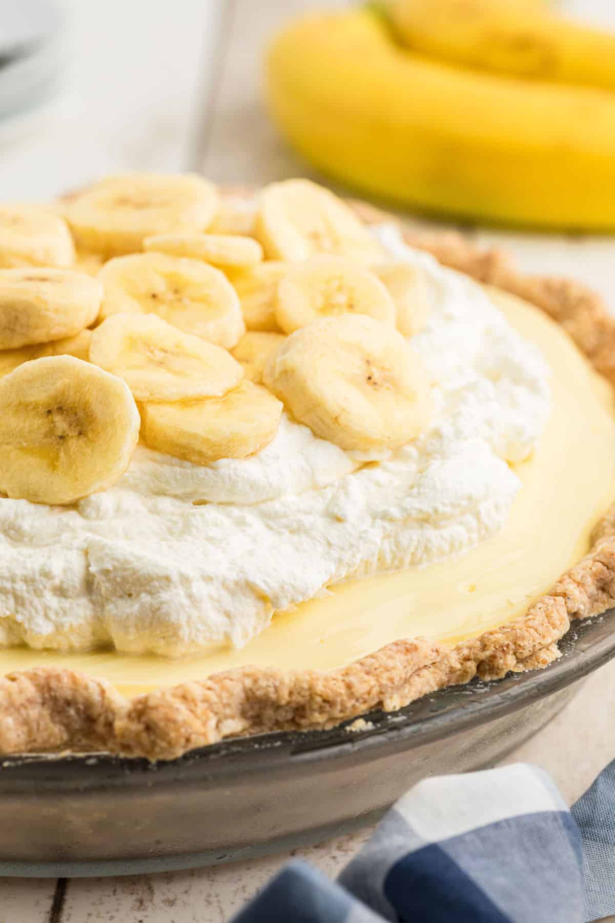 Side view of a dish full of banana cream pie, with some bananas in the background.