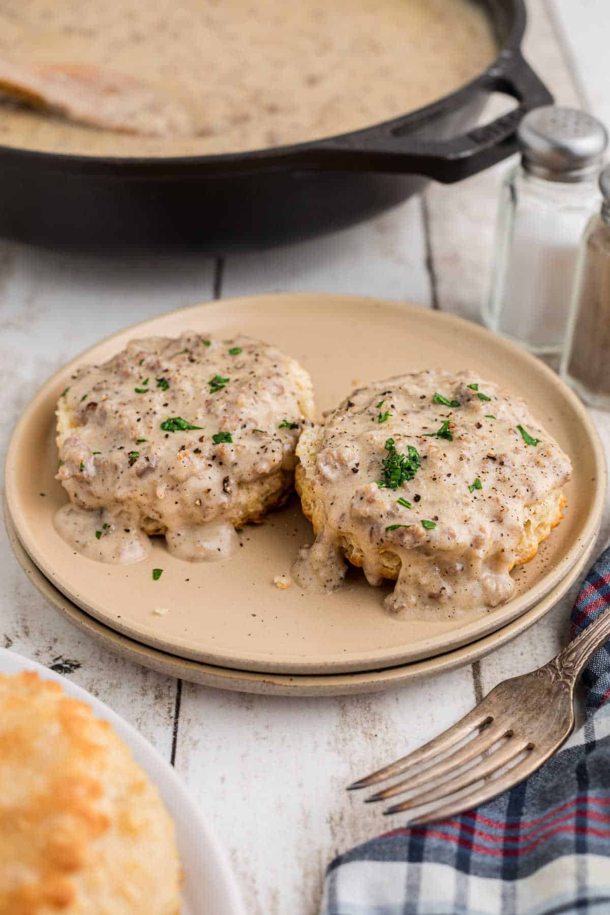 Biscuits and gravy on a plate with a skillet full of sausage gravy in the background.