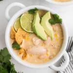 A square image of a bowl of white chicken chili with some avocado and limes and chips.