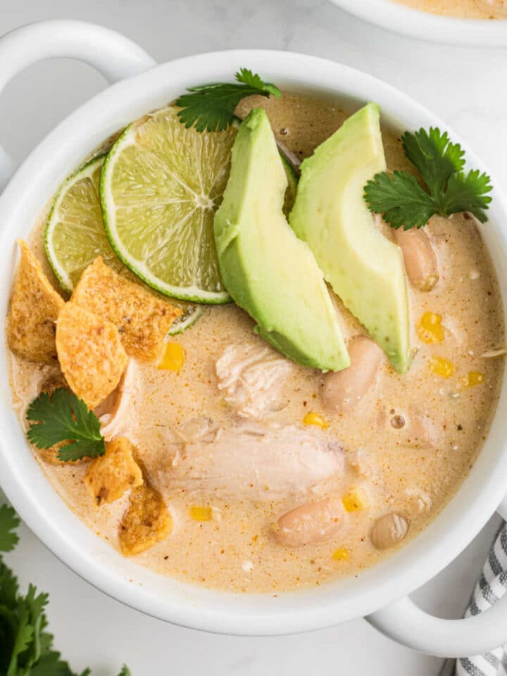 A square image of a bowl of white chicken chili with some avocado and limes and chips.