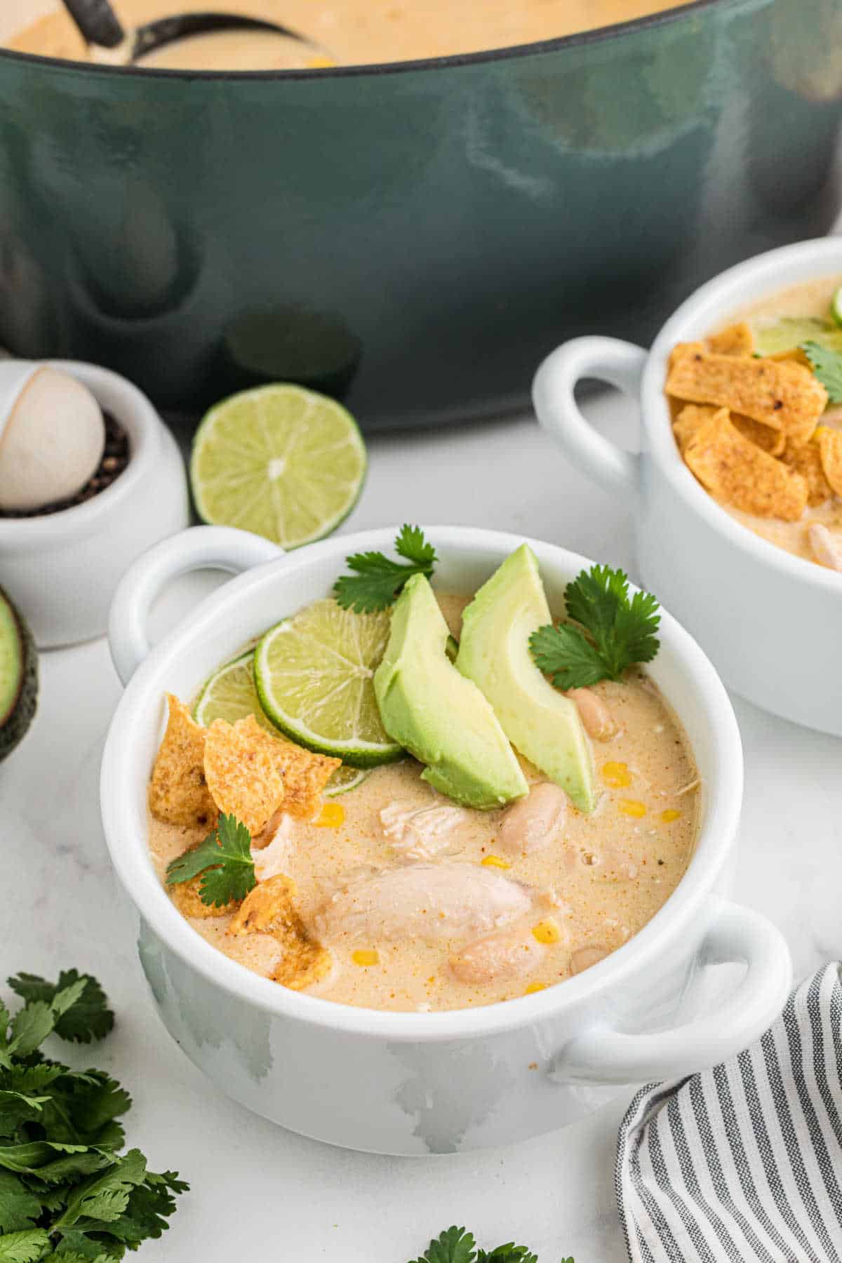 Two bowls of white chicken chili in front of a green pot. The chicken chili has some avocado pieces and chips on top.