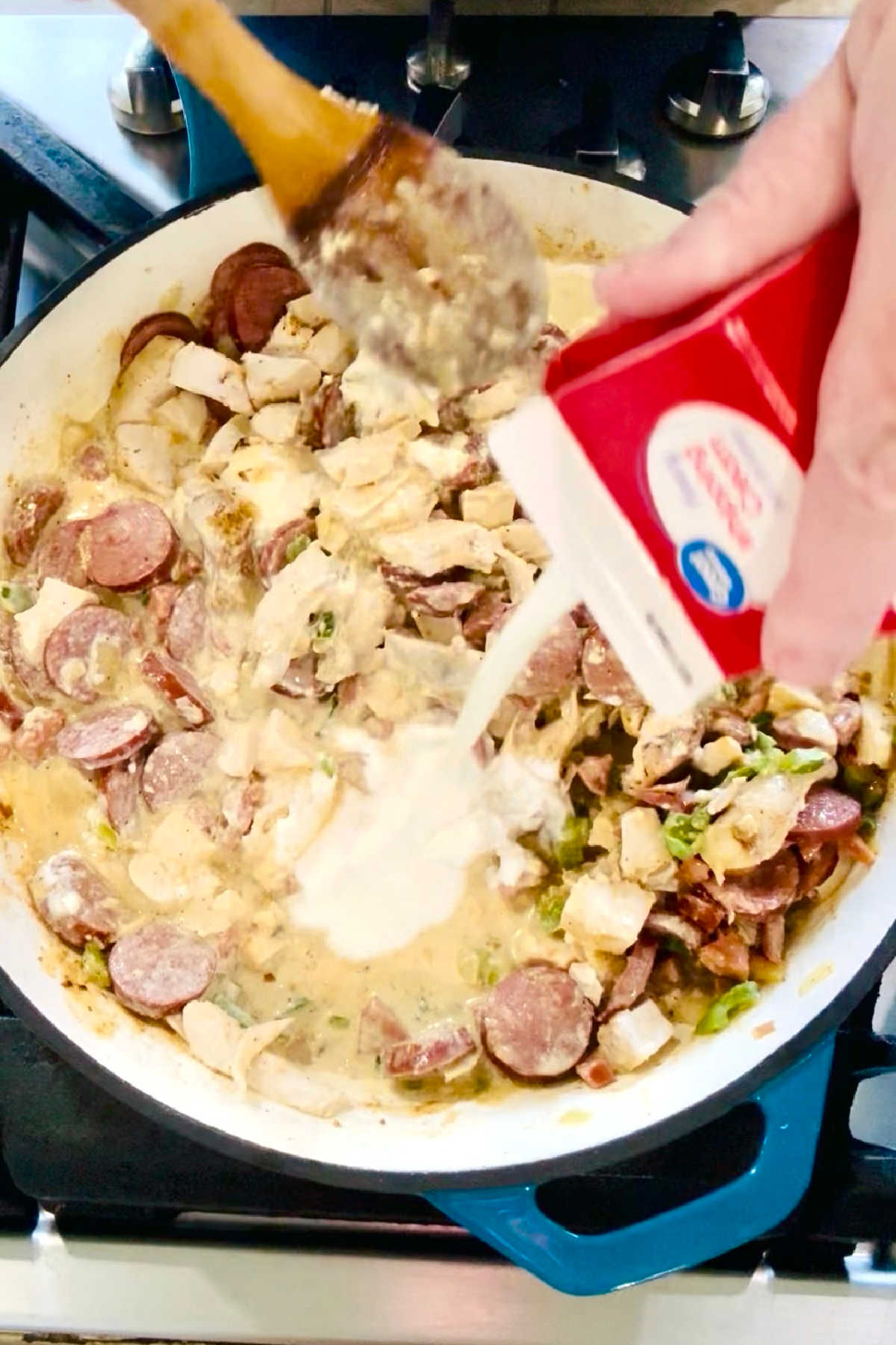 Cream being poured into a large skillet with chicken, sausage and cream cheese.