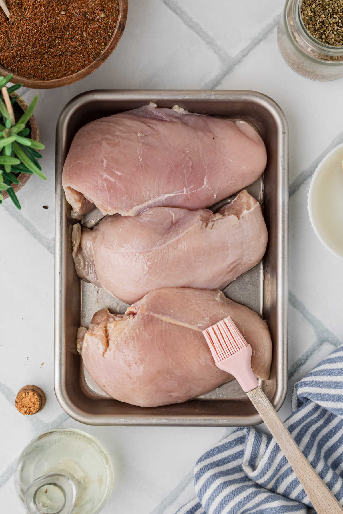 A pan with three chicken breasts, being brushed with oil.